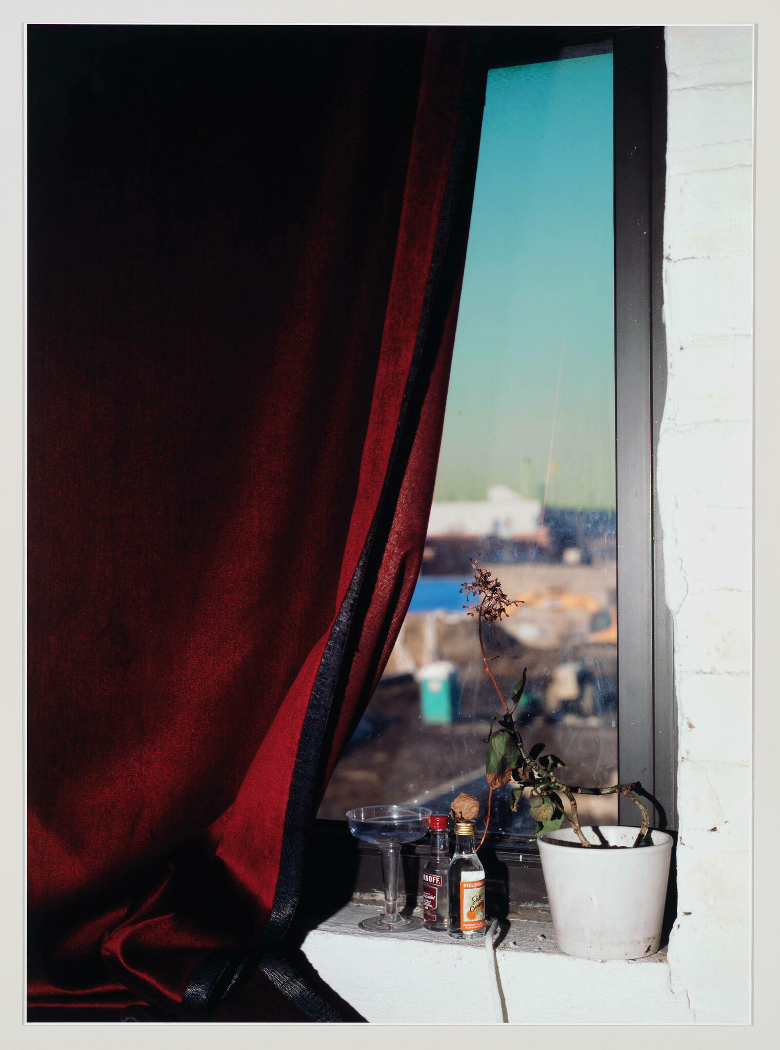 A richly colored red curtain with a black edge is partially drawn back to reveal a window sill. On the sill, there is a wilted plant in a white pot, a small bottle of alcohol, a miniature bottle of hot sauce, and an empty glass dish. The window provides a view of a bright blue sky and an urban landscape with buildings in the distance. The white-painted window frame contrasts with the dark curtain and the items on the sill.