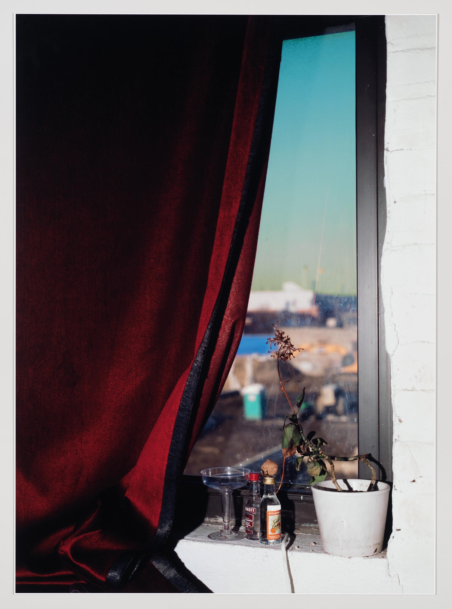 A red curtain partly covers a window ledge with a wilted potted plant, two small bottles, and a glass.