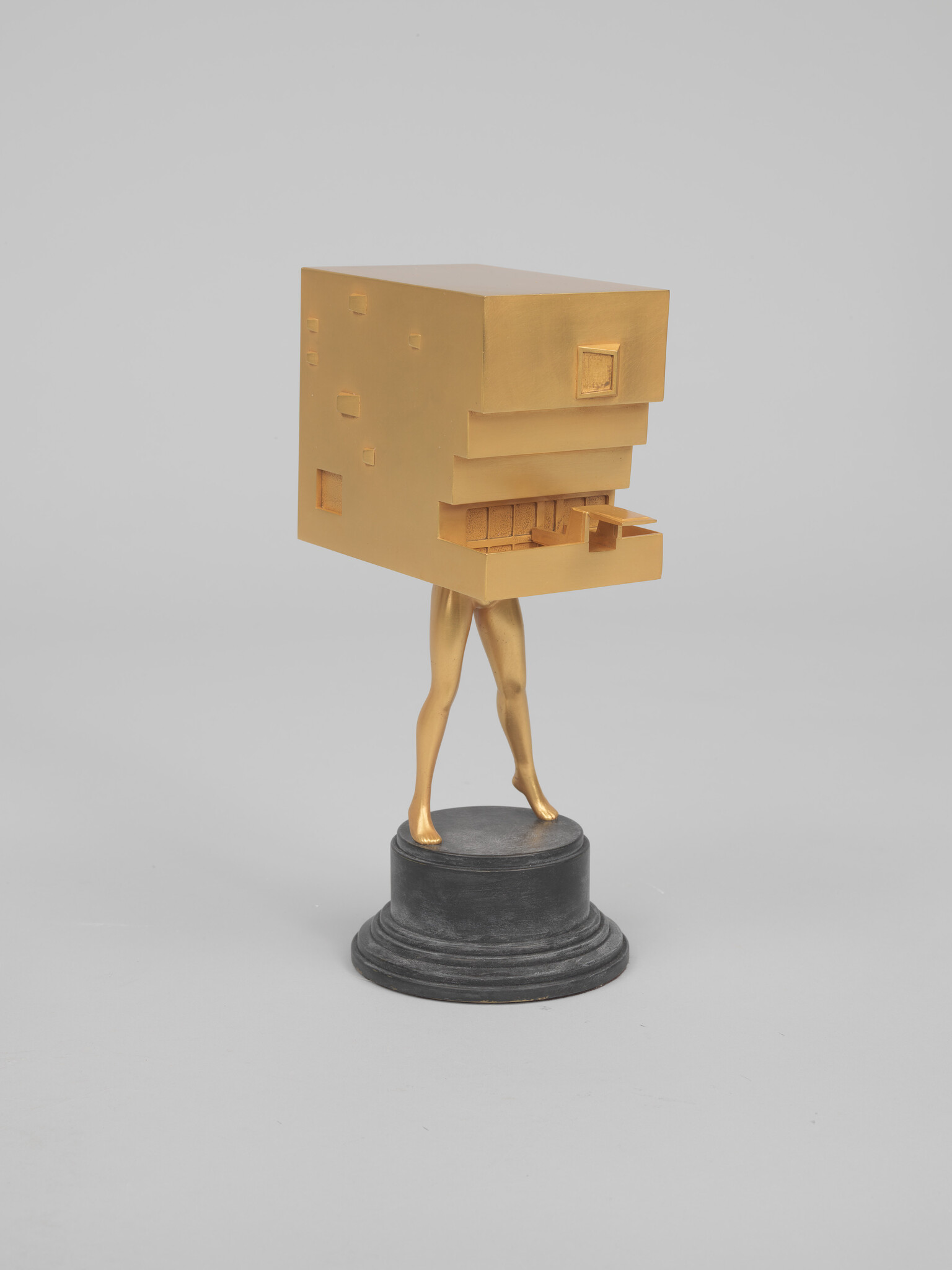 Golden sculpture showing a blocky head with stair-like mouth standing on human legs atop a black pedestal.