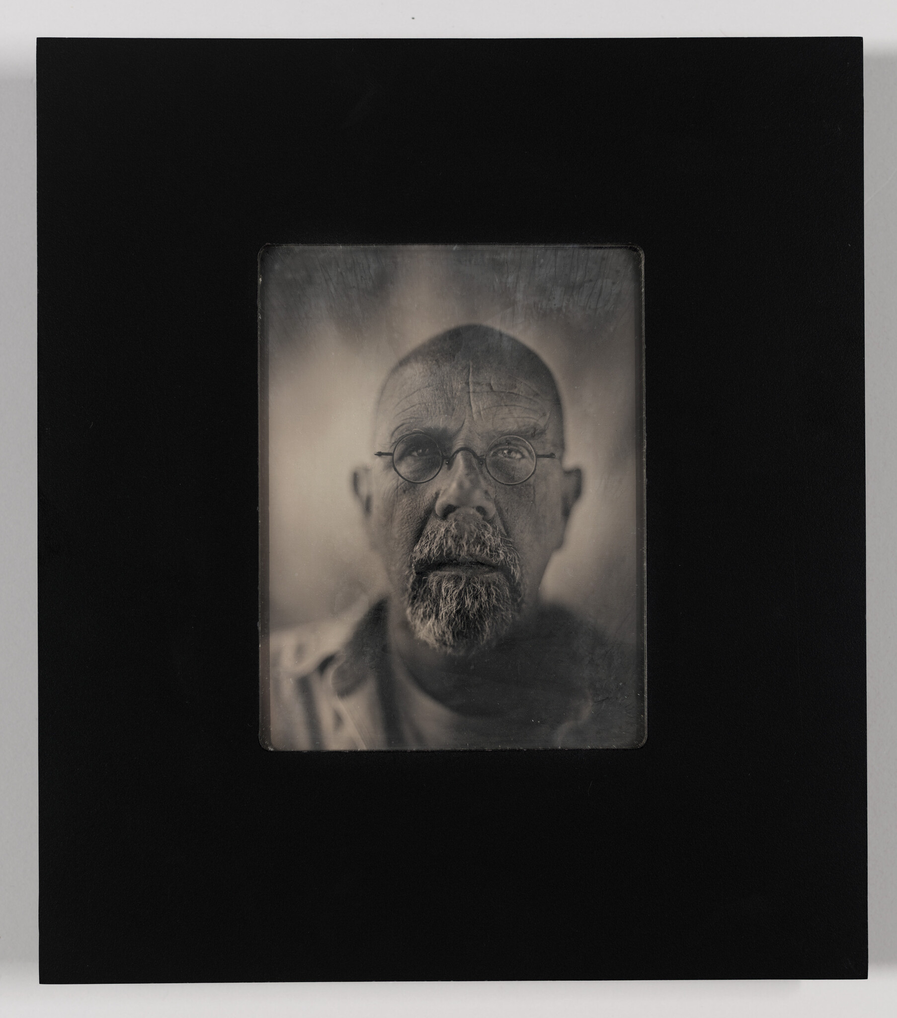 A monochromatic portrait of a bearded man with glasses, looking directly at the camera with a serious expression. The photo is centered within a square frame, set against a black background, giving it a vintage appearance.