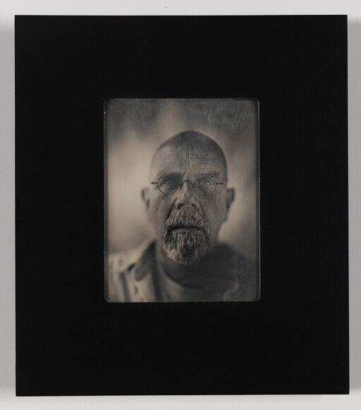 Bald man with round glasses and a beard stares straight at the camera in a mounted portrait.
