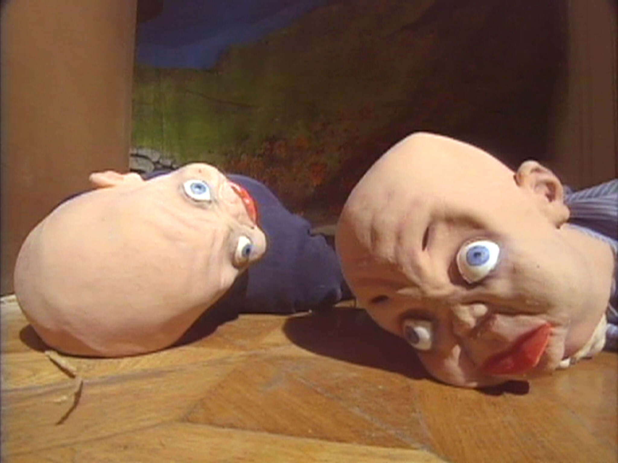 Two clay puppet heads with wide eyes and exaggerated features lying on a wooden floor, facing sideways.