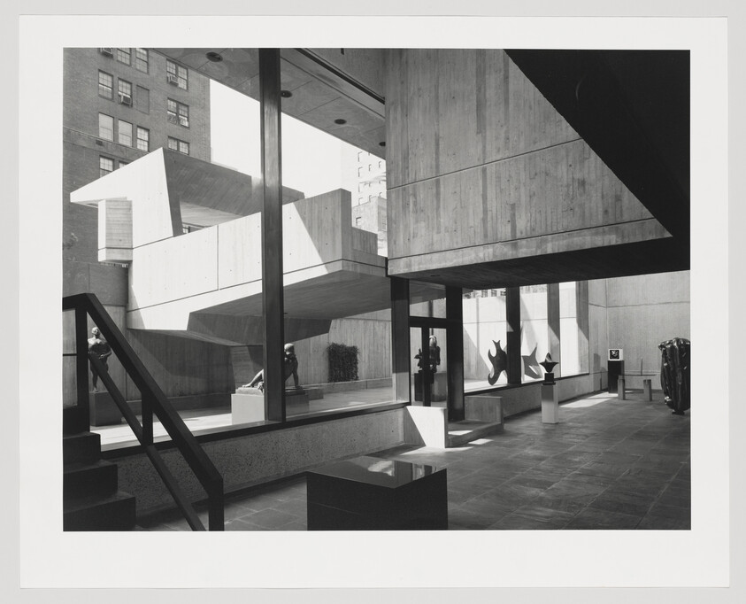 Museum lobby with concrete architecture and abstract sculptures visible through large glass windows.