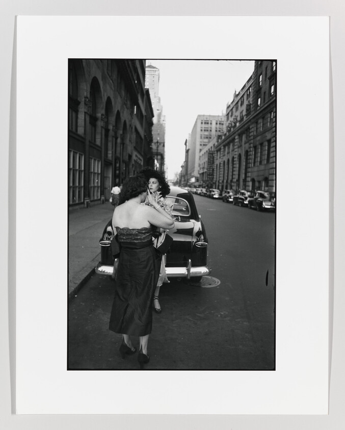Two women in dresses standing by a parked car on a city street, one fixing her hair.