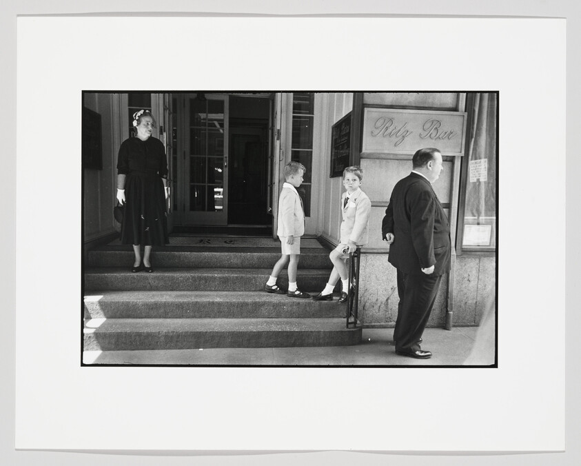 Two boys in short suits stand on hotel steps near a sign reading "Ritz Bar" while adults watch.