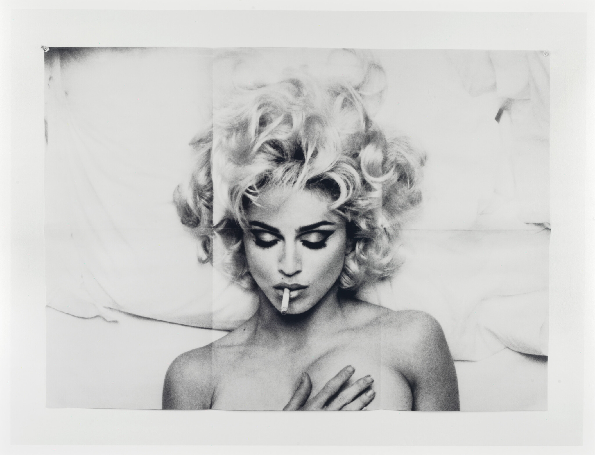 Blonde woman with curly hair lies against pillows, holding a cigarette in her mouth and hand on chest.