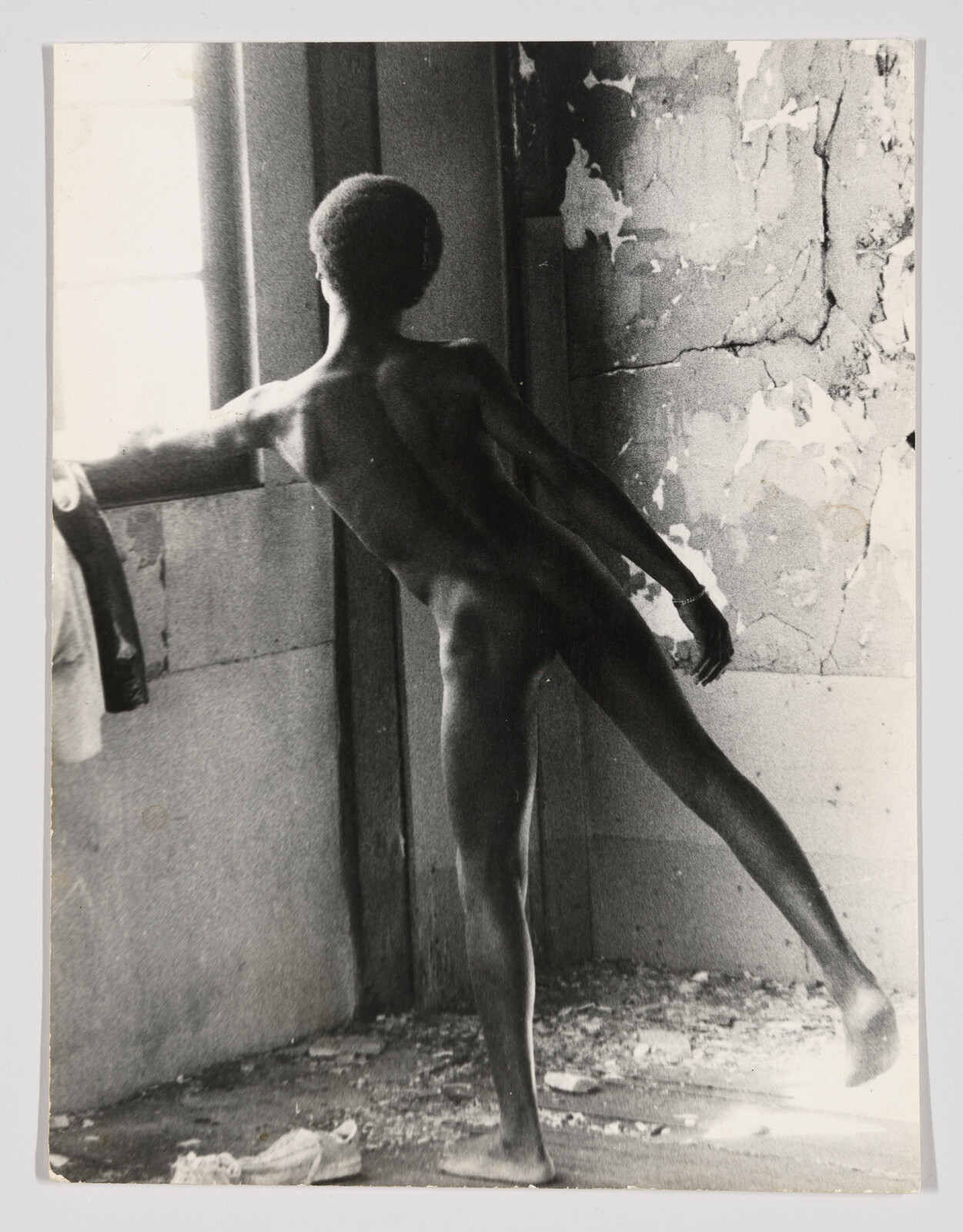 A naked Black man balances on one leg while looking out into the glistening light from a window in a dilapidated room