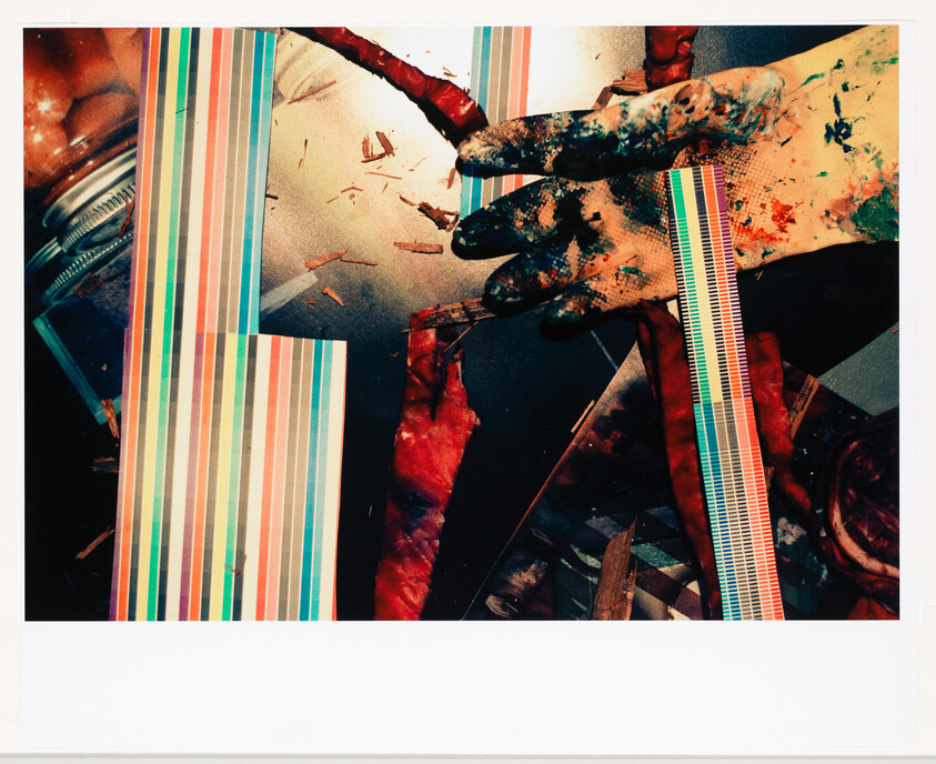 Paint-covered gloved hand reaches across a table cluttered with striped color samples and debris.