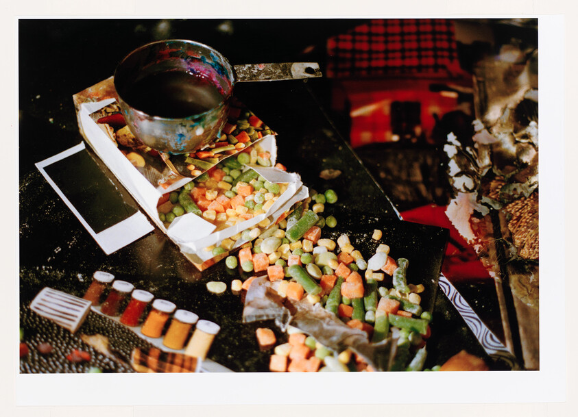 A torn box of mixed vegetables spilled across a cluttered table next to a metal saucepan.