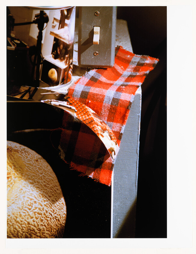 Red plaid cloth draped over a gray shelf edge beneath a small wall switch, with a cantaloupe nearby.