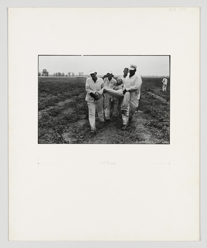 Five men carry an injured worker across a muddy field while others watch in the distance.