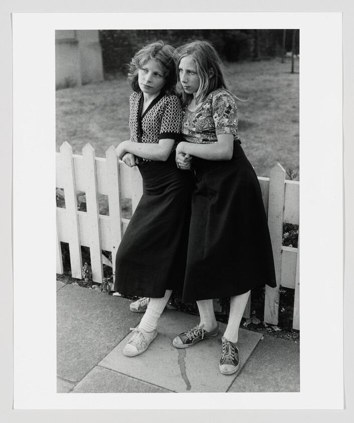 Two white women, wearing patterned blouses and a long maxi skirt, lean their hips aganist each other and curl up their fists