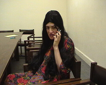 Woman with long dark hair in a floral dress sits at a table and speaks on a cellphone.
