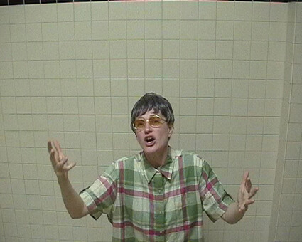 Person wearing glasses and a plaid shirt gestures expressively with both hands in front of a tiled wall.