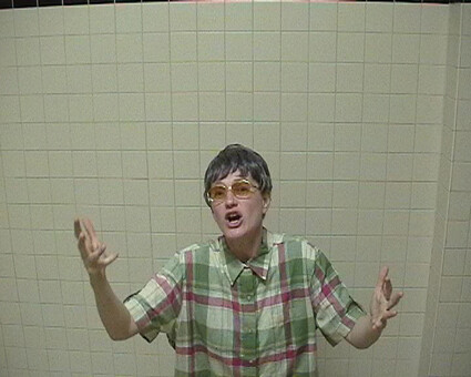 Person wearing glasses and a green plaid shirt gestures with both hands against a tiled wall.