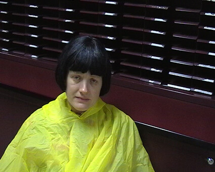 A person wearing a bright yellow poncho sits indoors against a dark wooden wall with blinds.