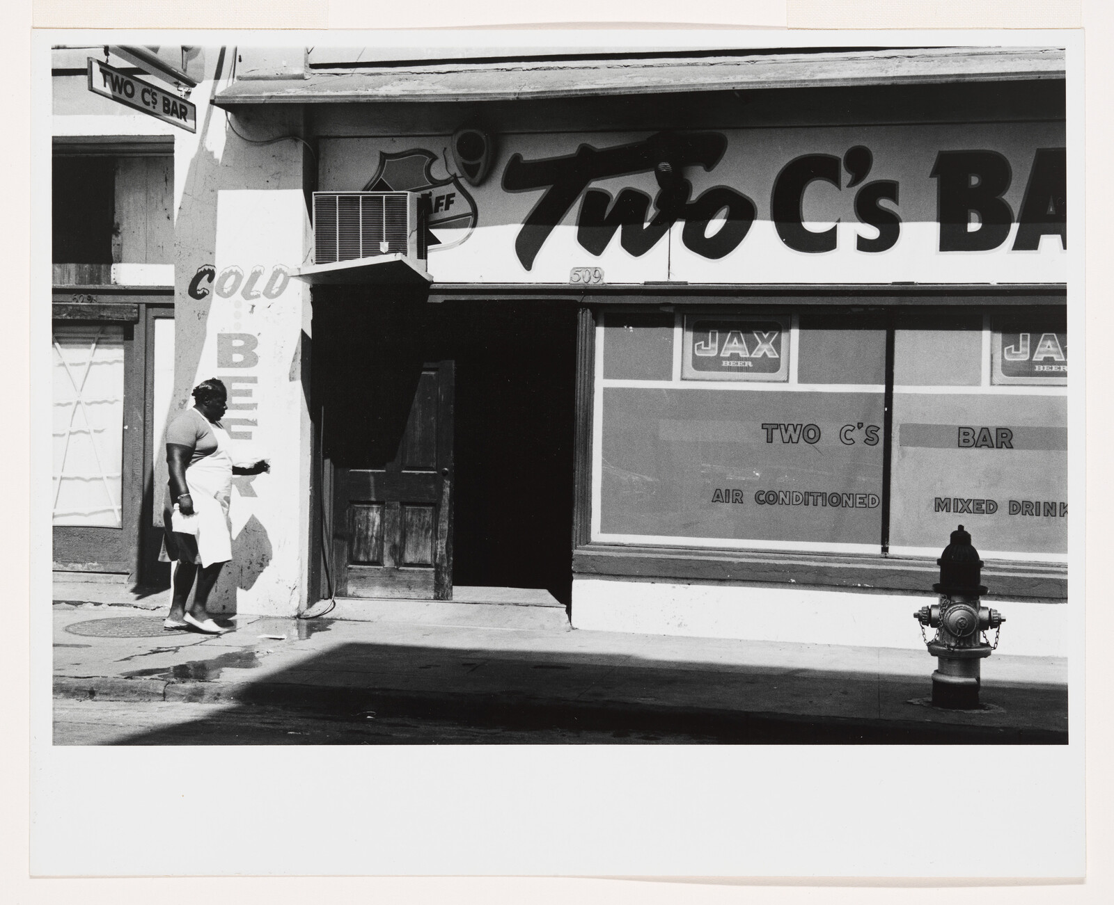 A woman in an apron walks past the entrance of Two C's Bar on a sunny street.