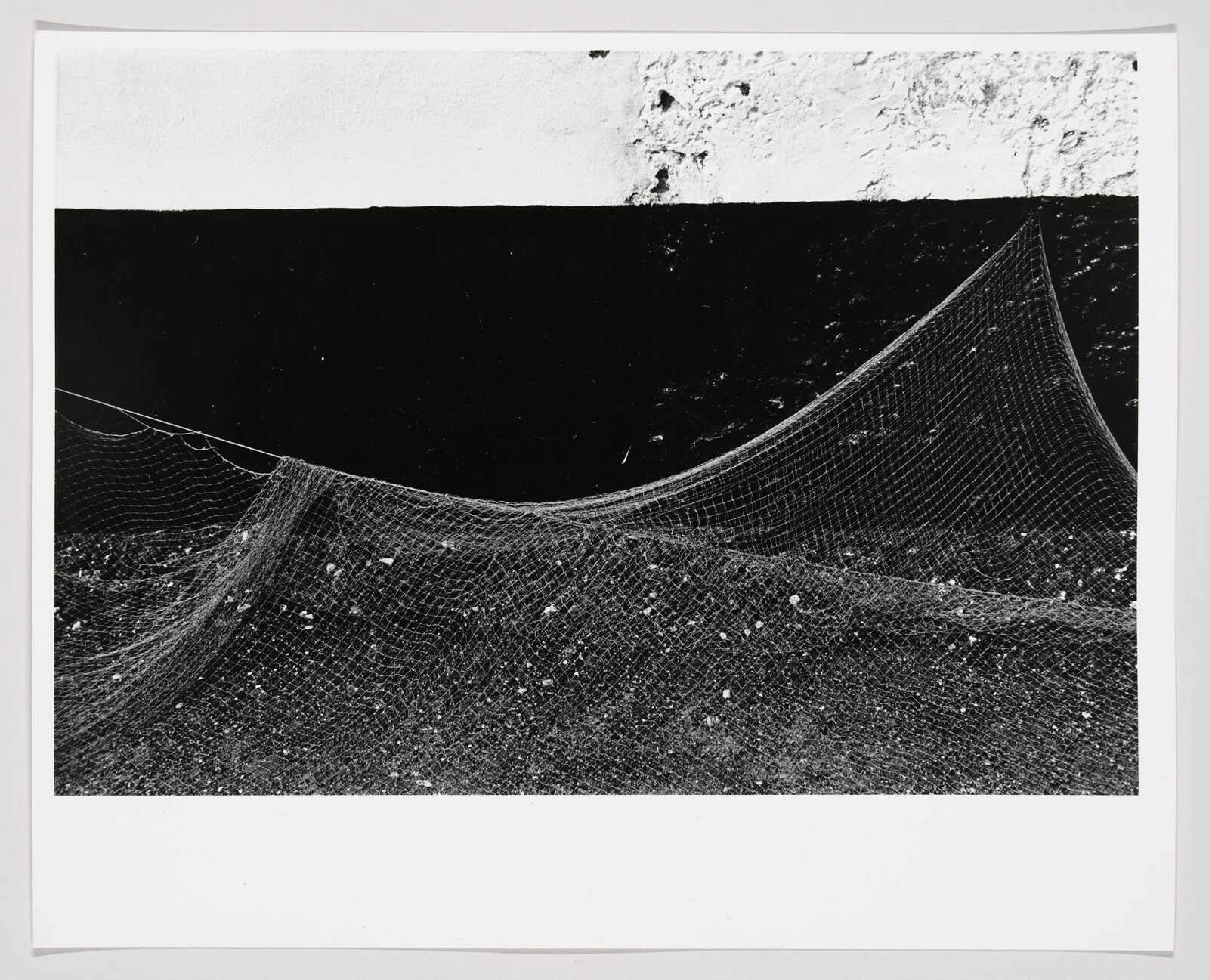 A large fishing net draped over pebbly ground, its mesh curving against a dark wall.