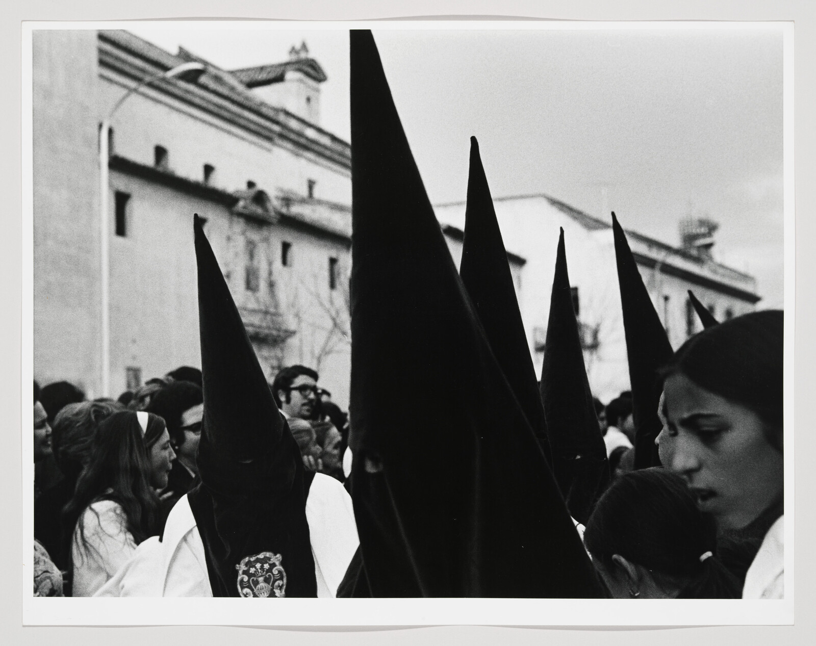 Hooded participants in tall pointed hats march through a crowded street during a religious procession.