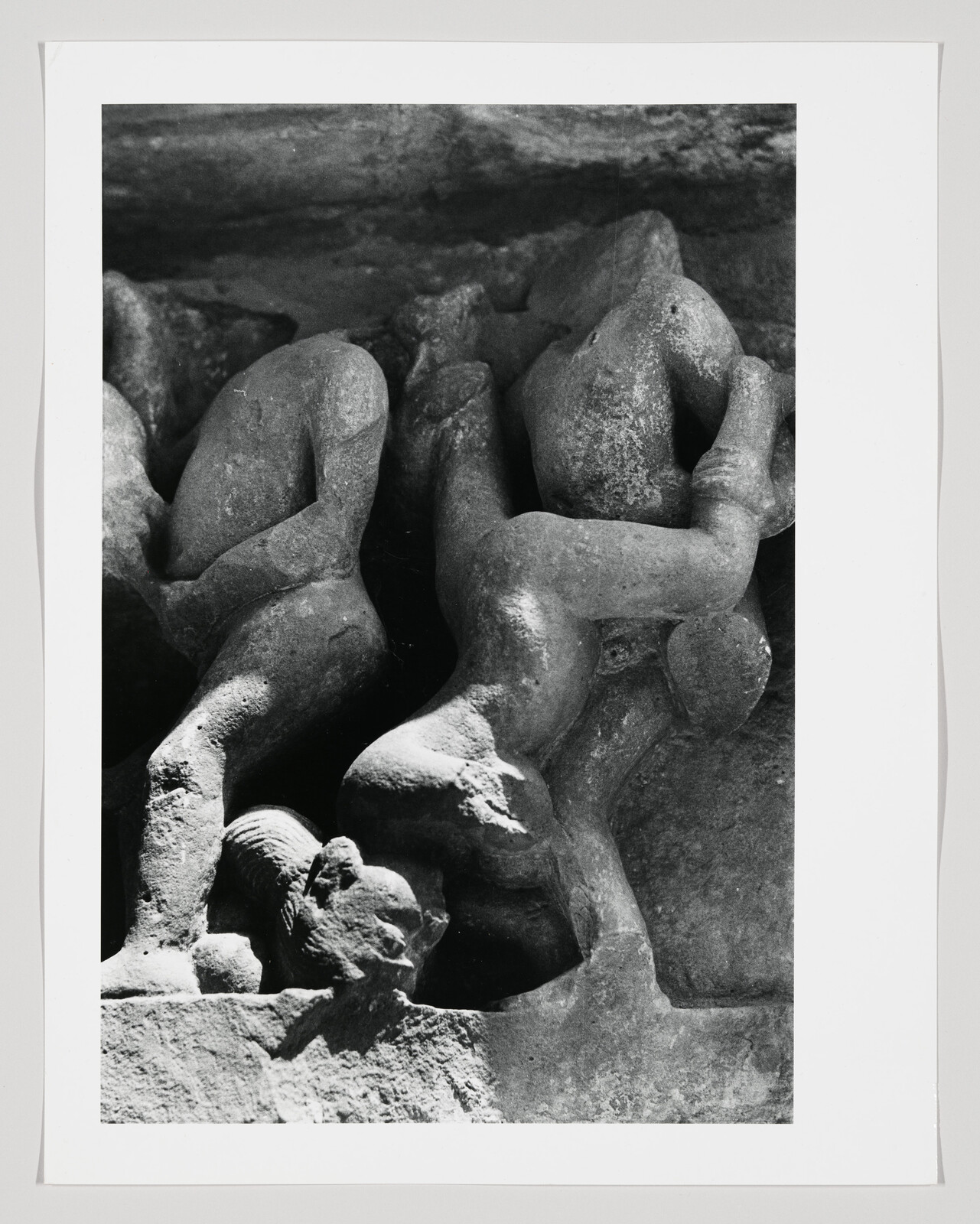 Two weathered stone figures entwined on a carved relief with a small animal head at their feet.