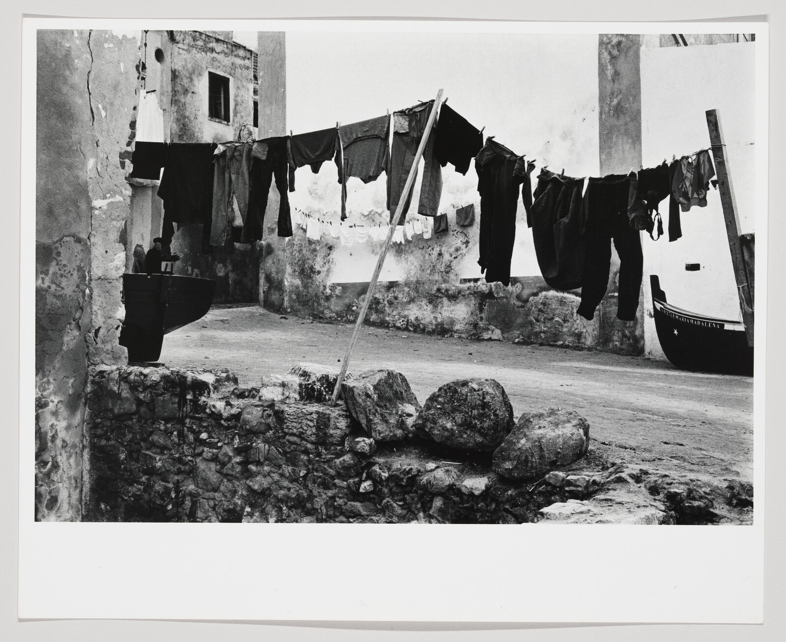 A black and white photograph depicting a rustic outdoor scene with clothes hanging on a line to dry between dilapidated buildings. In the foreground, there are large stones and a washbasin, and to the right, a boat is visible with the name "Nossa Senhora da Atalaia" on its side. The setting appears to be a traditional courtyard or alleyway, evoking a sense of daily life in a bygone era.