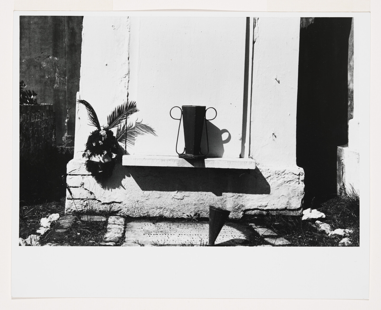Black and white photograph showing a rustic outdoor scene with a watering can placed on a stone step next to a potted plant with large fronds. The strong sunlight casts sharp shadows of the objects onto the wall and ground, highlighting their contours. The background features the textured wall of a building with a visible corner edge, and the ground appears to be a mix of dirt and sparse grass.