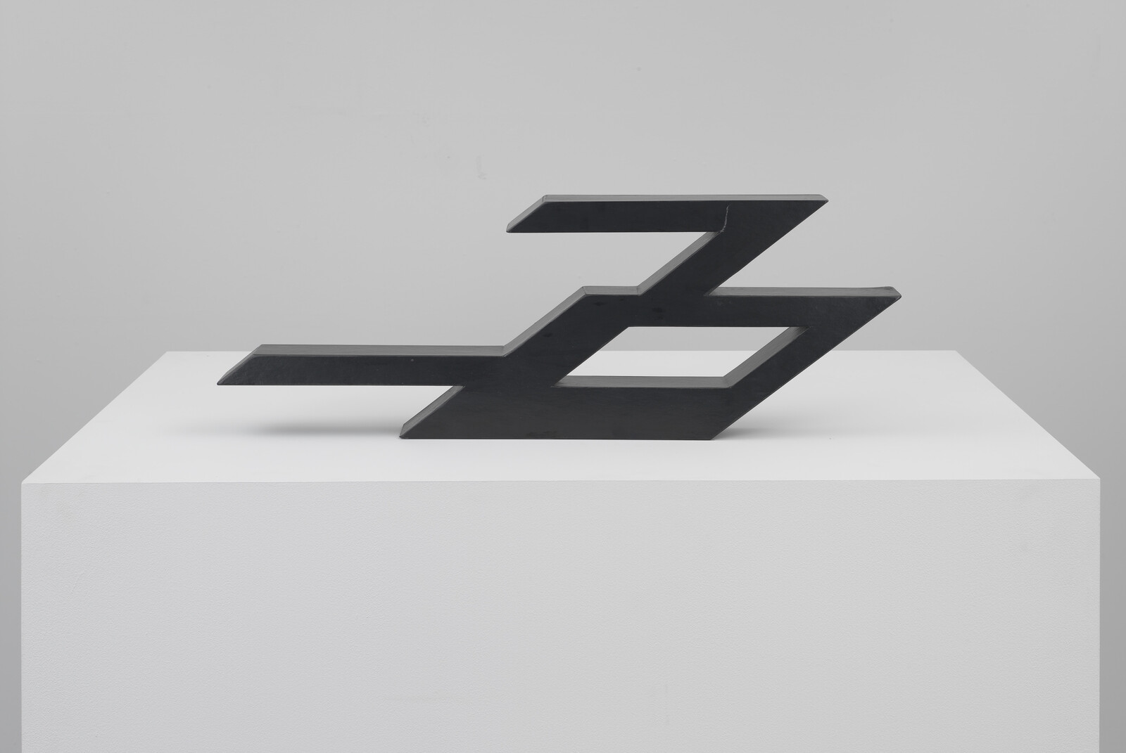 A black abstract sculpture with geometric shapes resembling a stylized number three, displayed on a white pedestal against a light grey background.