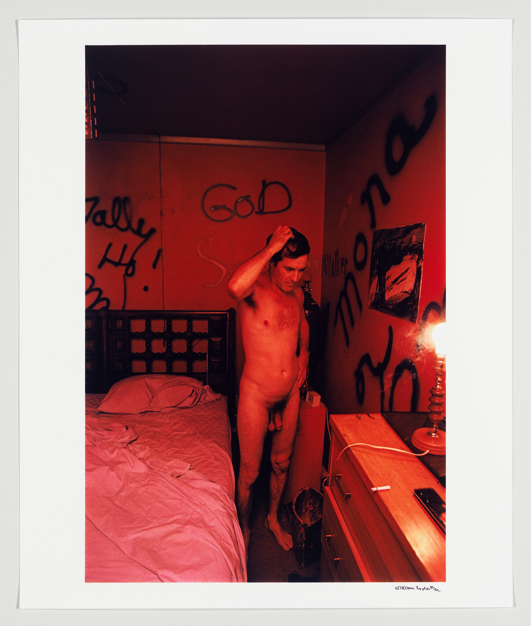 A photograph of a room with red lighting, featuring a nude man standing beside a bed with his hand on his head. The walls are covered in graffiti with words like "God" and "Sally." A candlestick with a lit candle is on a dresser, contributing to the room's illumination.