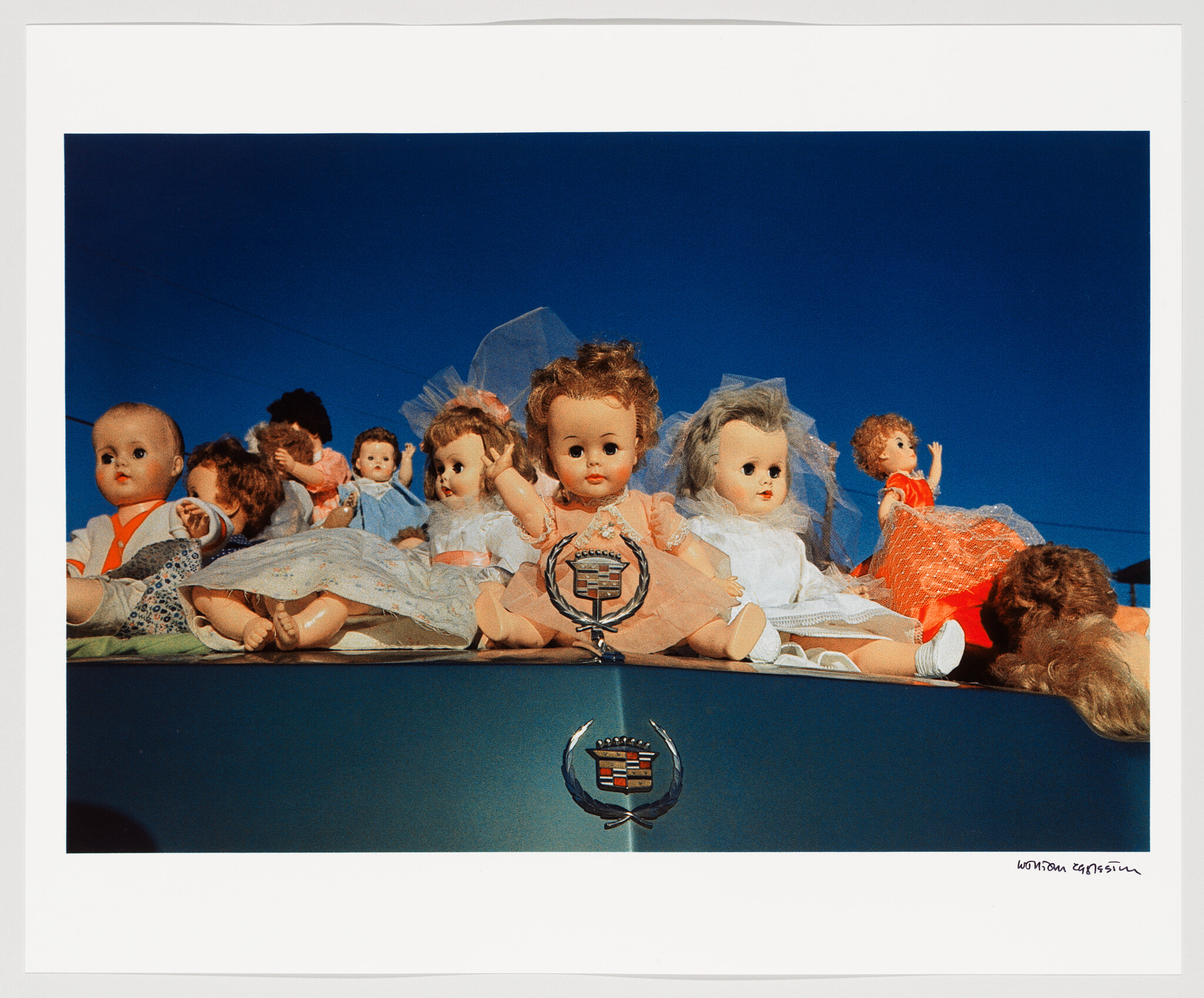 Several vintage dolls in dresses sit on a car hood above a Cadillac emblem.