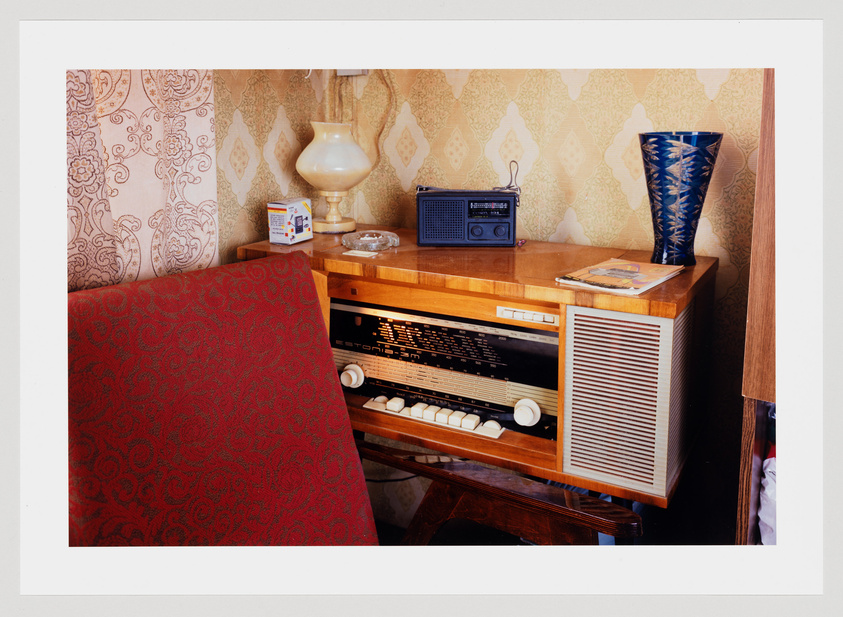 A vintage interior scene with a patterned wallpaper background and a wooden sideboard hosting a classic radio, a lamp, and a blue vase. To the left, a portion of a red patterned chair is visible, and on the sideboard, there's a small portable radio, an ashtray with cigarette butts, a pack of cigarettes, and a book. The atmosphere suggests a mid-20th-century setting.