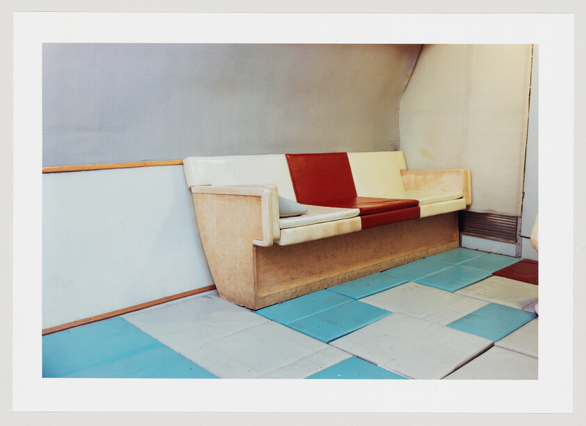 A worn three-seat bench with red and white cushions against a pale wall over blue tiles.