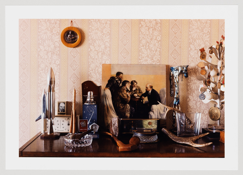 A well-arranged collection of personal items and memorabilia on a wooden surface against a wallpapered wall with a floral pattern. The collection includes a clock, a rocket-shaped pen holder, a framed photograph, perfume bottles, a crystal ashtray, and various decorative objects. A large framed painting of a group of people in conversation is leaning against the wall, partially overlapping with a mirror and a tree-shaped jewelry holder adorned with necklaces and trinkets. To the right, a pair of antlers lies in front of a glass vase with a unique melted design. A small round portrait hangs on the wall, and the scene is bordered by a white matting within a frame.