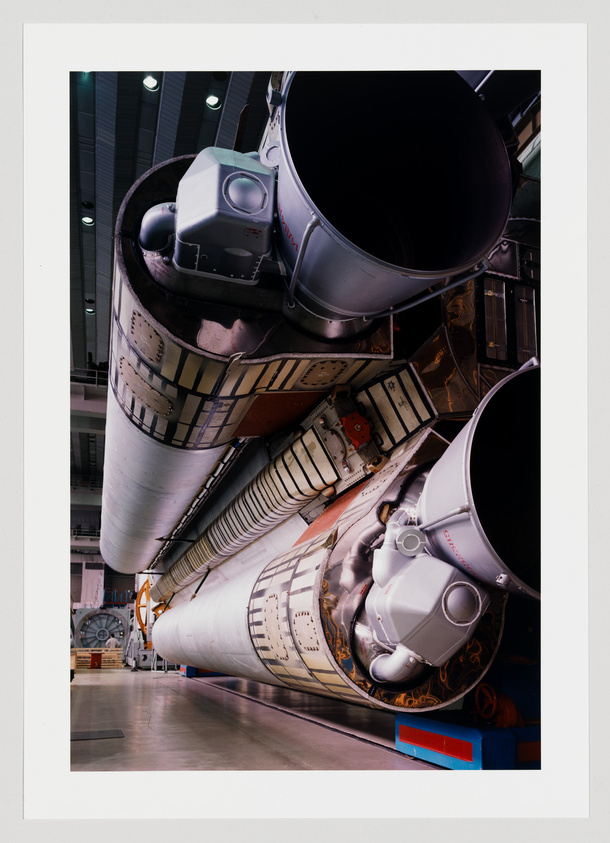 A close-up view of the side of a space shuttle, showcasing the detail of its white external tank and two of its main engines with nozzles, inside a hangar with industrial lighting.