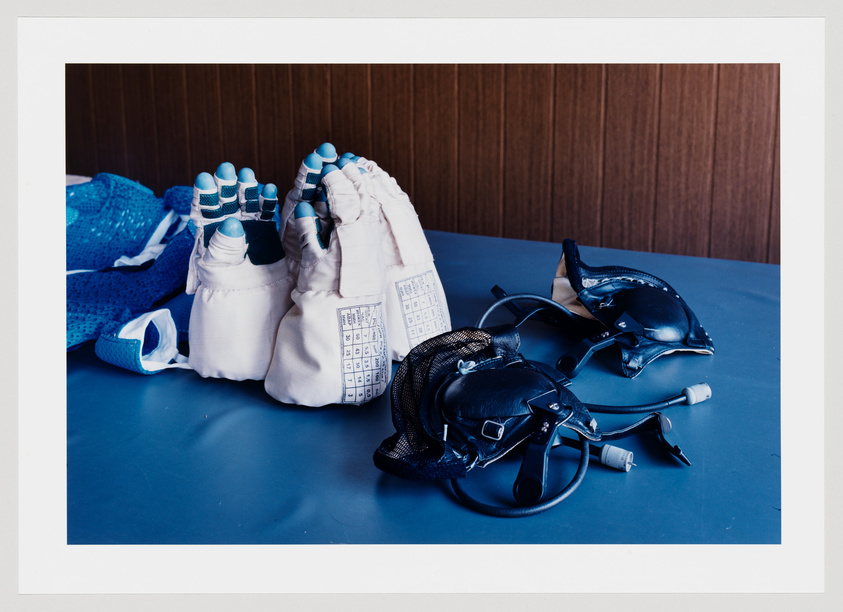 A collection of fencing gear laid out on a blue surface, including a white glove with blue padded fingertips, a mesh mask, and a black body cord. The background features a wooden panel.