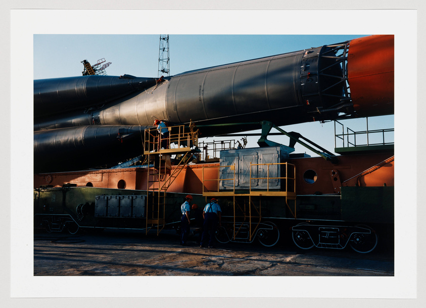 A large, segmented rocket with a black body and red nose cone is being transported on a multi-wheeled launch vehicle. Workers in hard hats are visible around the base, with some on the ground and others on elevated platforms attached to the rocket. The scene is set against a clear sky with industrial structures in the background.