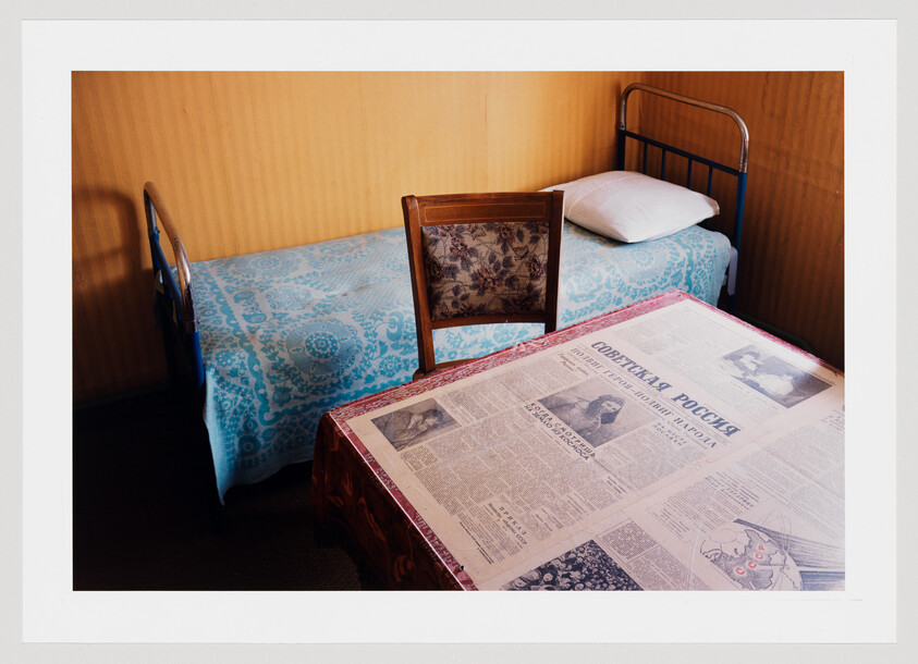 A simple room with a single bed, wooden chair, and table covered with a Soviet-era newspaper.