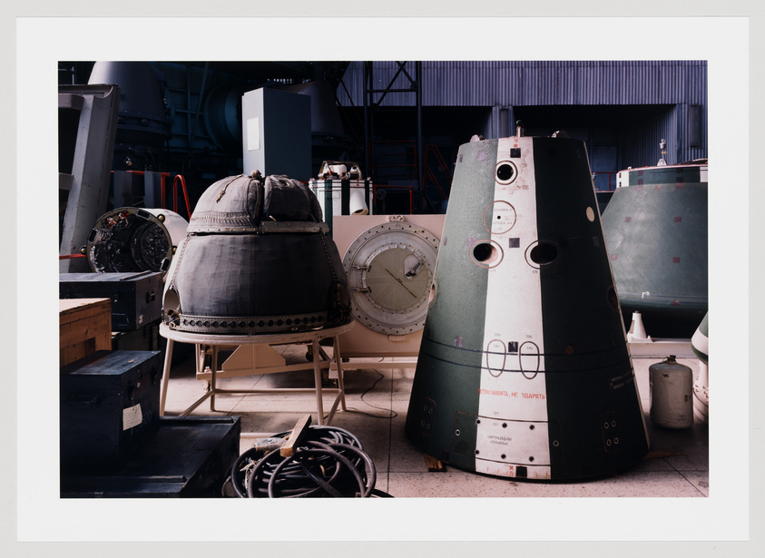 A photograph of various spacecraft components and modules in a workshop setting, including a partially disassembled capsule with its heat shield visible, and other conical capsule sections with Cyrillic script, suggesting Russian spacecraft. The environment is industrial with tools and parts scattered around, and the overall scene conveys a sense of space exploration technology in maintenance or assembly.