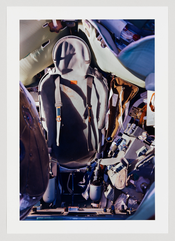 A detailed view of an ejector seat from a military aircraft, showing the seat's harnesses, buckles, and survival equipment. The cockpit around the seat is filled with various controls and instruments. The image is framed with a white border.