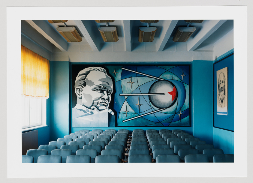 A photograph of an auditorium with rows of empty grey seats facing a stage with a large mural. The mural features a monochromatic portrait of a man on the left and an abstract design with a red arrow on the right. The room has a blue color scheme with a single yellow curtain on a window to the left.