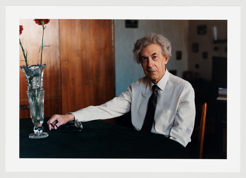 Elderly man in a white shirt and tie sitting at a table with a vase, holding glasses.