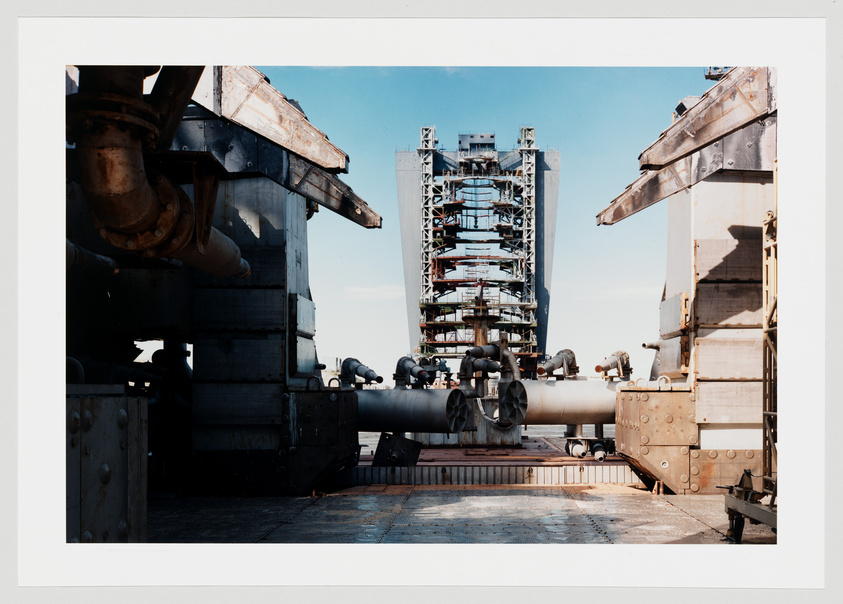 A photograph showing a large industrial structure with a complex array of pipes and scaffolding, flanked by heavy machinery parts, under a clear blue sky.