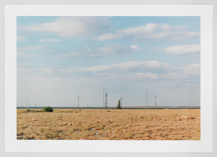 A wide-open landscape with dry grassland under a partly cloudy sky, featuring several tall, narrow metallic structures, possibly part of a rocket launch site, in the distance.