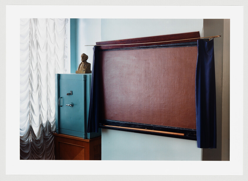 An empty pull-down brown chalkboard with side curtains next to a teal safe and bust.