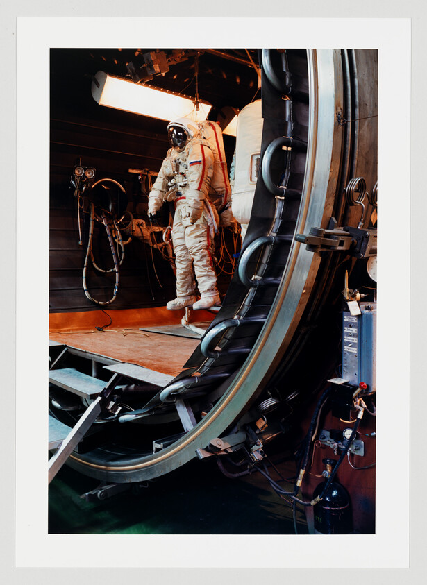 An astronaut wearing a space suit stands inside a large circular centrifuge training chamber.