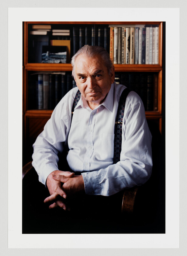Older man in a white shirt and suspenders sits with hands clasped in front of a bookshelf.