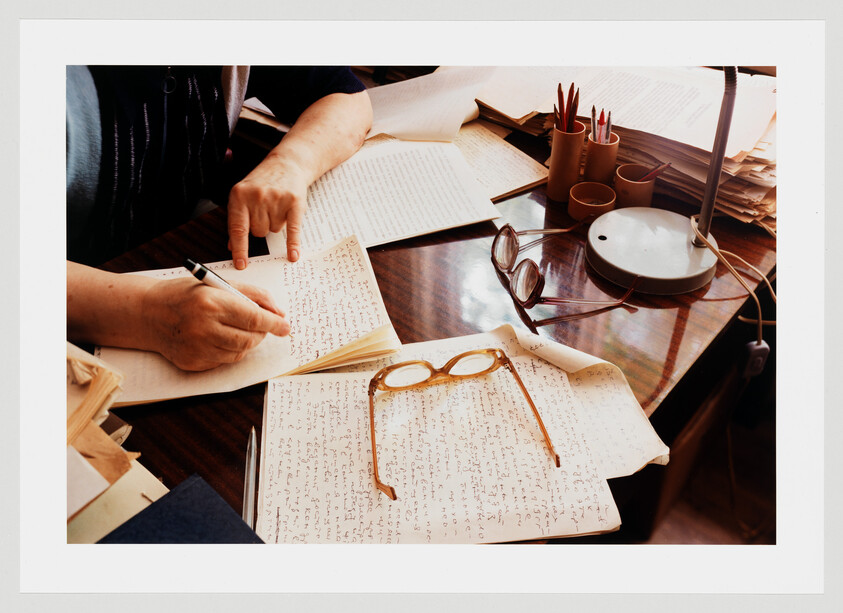Hands writing notes at a cluttered desk with eyeglasses resting on handwritten pages.