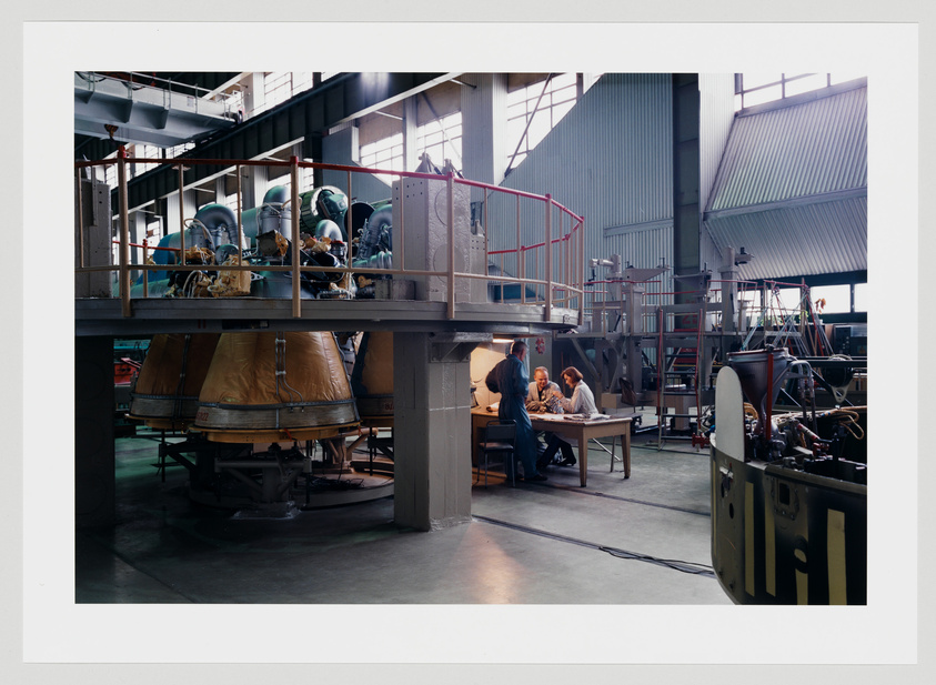 A spacious industrial facility with high ceilings and large windows casting natural light onto the floor. In the foreground, there are large pieces of machinery, possibly parts of rockets or engines, covered with protective tarps. On an elevated platform in the middle, several people in work attire are gathered around a table, seemingly engaged in a discussion or review of documents. The background is filled with various industrial equipment and structures, suggesting an active engineering or manufacturing environment.