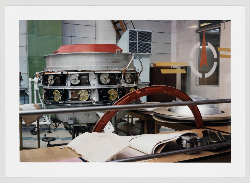 A spacecraft module sits on a workbench surrounded by tools, plans, and assembly parts.