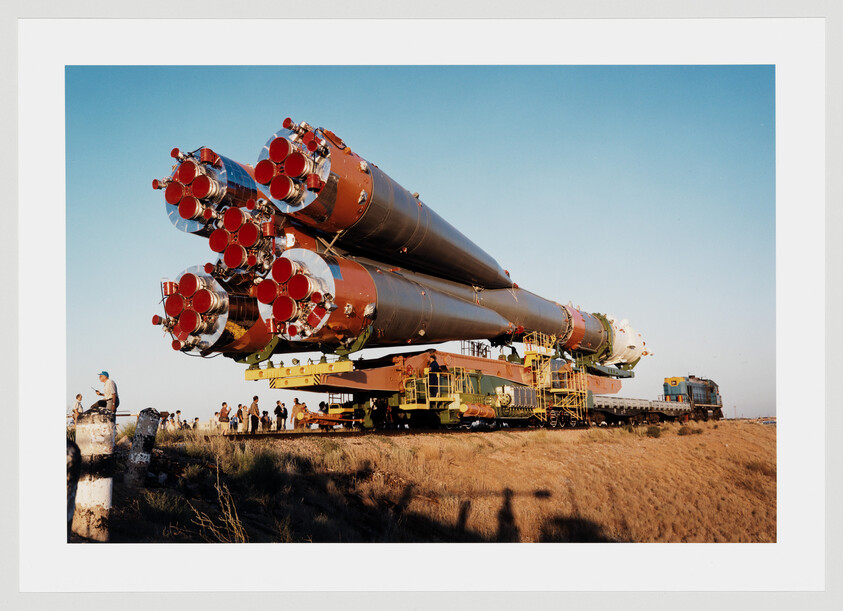 A large multi-stage rocket is being transported on a flatbed train with workers nearby.