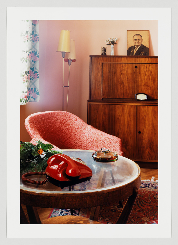 A cozy vintage room featuring a red textured armchair, a round wooden table with a red rotary phone and an ashtray on top, a wooden cabinet with a framed portrait and a small vase of flowers, a standing lamp with a yellow shade, and floral curtains. The room has a warm ambiance with a patterned rug on the floor.